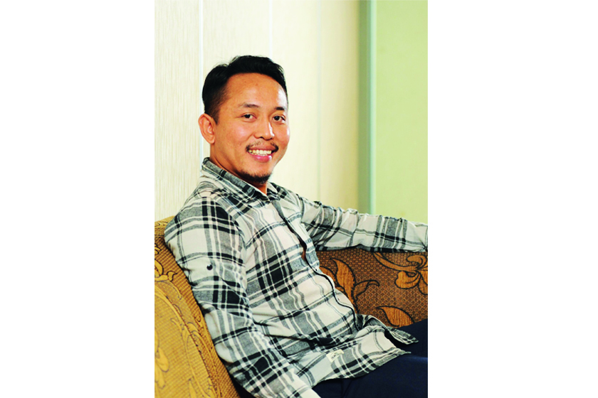 A man in a squared patterned t-shirt smiles and sits on the sofa